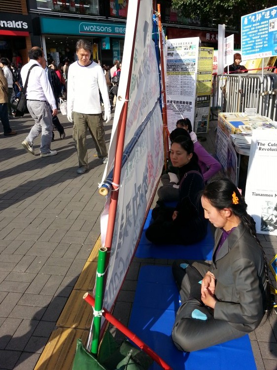 Practicanţii Falun Gong sunt văzuţi meditând la punctul de informare de la terminalul de feribot din Hong Kong, la 13 februarie 2013. Un suport de banere ridicat ulterior de Asociaţia de Îngrijire a Tineretului din Hong Kong blochează trecătorii să vadă punctul de informare al practicanţilor. Şirul de dale galbene construit la baza banerelor asociaţiei constituie o potecă destinată orbilor.