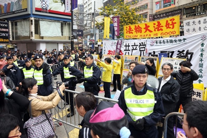 Practicanţii Falun Gong demonstrează unul dintre exerciţiile disciplinei la un loc de informare de la Causeway Bay din Hong Kong, în timp ce poliţia din Hong Kong îi protejează de membrii îmracaţi în tricouri verzi ai Asociaţiei de Îngrijire a Tinererului din Hong Kong, la 3 martie, în apropiere de departamentul magazinului Sogo.