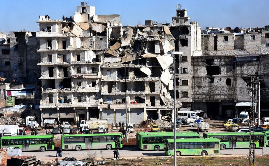 Autobuze folosite pentru evacuări aşteaptă în faţa unor clădiri distruse din oraşul sirian Alep.