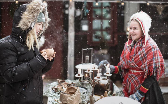 Două fete stau la o masă şi servesc un "fika" în aer liber, în zăpadă. Pe masă se află un candelabru vechi cu lumânări aprinse. Una dintre fete poartă un costum Sámi.